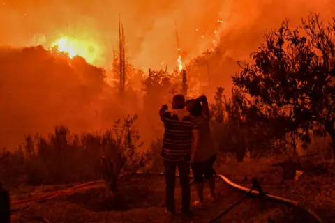 VALERIE GACHE/AFP A couple look on at a wildfire raging in the village of Ano Loutro, south of Athens, on 30 September, 2024.