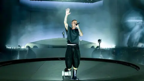 Getty Images Justin Bieber is wearing a grey-black t-shirt and cropped black baggy jeans. He is singing into a handheld mic with one arm raised. He has a shaved head, tattoos covering both arms and his neck and a shaved head. He is wearing black boots standing on a circular stage with a dark backdrop. 