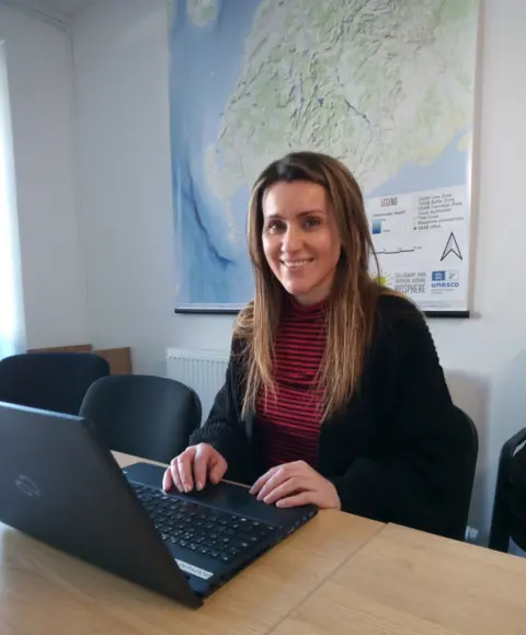 A woman with long brown hair in a red jumper and black cardigan sits at a laptop on a wooden desk with a map in the background