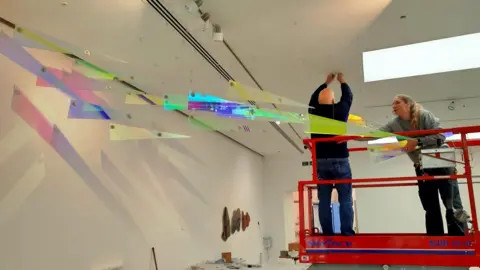 Two technicians, a man and a woman, on a red hydraulic platform in a white museum room. They are putting the final pieces to a glass artwork which hangs from the ceiling. It is a series of glass triangles in different colours which reflect on to the white walls. 