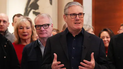 Prime Minister Sir Keir Starmer dressed in a dark jacket and dark blue shirt with grey hair and glasses is stood in front of a crowd of people. Behind him also is the Mayor of the West Midlands Richard Parker, also in a dark jacket and a light blue shirt. He's wearing thick black rimmed glasses and has short white hair.