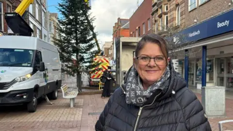 A woman smiling as she stands on Rhyl High Street in front of the tree. She is wearing a black padded coat and glasses.