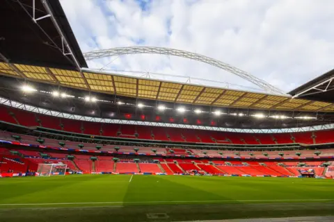 Tim Clayton/Getty Images An interior view of an empty Wembley Stadium. Thousands of red seats are visible next to a green pitch beneath a large arching roof structure and cloudy sky.