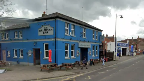 Google The Salopian bar is painted blue, and has wooden benches outside it. It it located next to a road. 