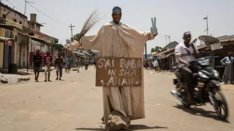 AFP via Getty Images A makeshift figure of Buhari, bearing the words "Sai Baba Inshallah", stands in the middle of a dusty road. A motorbike zooms past.