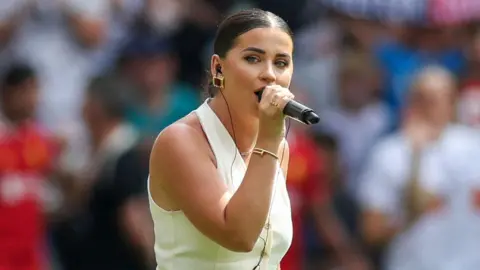 Claudia Rose singing before the match. She is wearing a cream halter neck waistcoat and has dark hair with a central parting tied back in a bun and chunky gold earrings. The crowd is blurred in the background