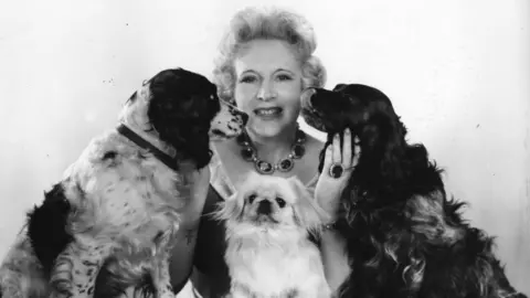 Cartland Promotions Barbara pictured in black and white with 3 dogs. She had coiffed wavy fair hair,shaped eyebrows and a chunky necklace and matching ring. She looks very glamorous.