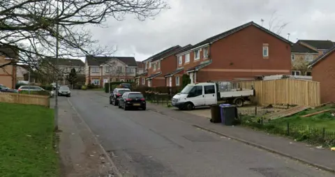 Residential street with brick semi-detached houses, parked cars along the road, a white van in a driveway, wooden fencing, and an overcast sky