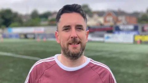 Jordon Connolly, con cabello negro y barba, lleva una camiseta blanca y rosa. Está detenido en una trama 3G, con mesas publicitarias y una posición de gol visible detrás de él.