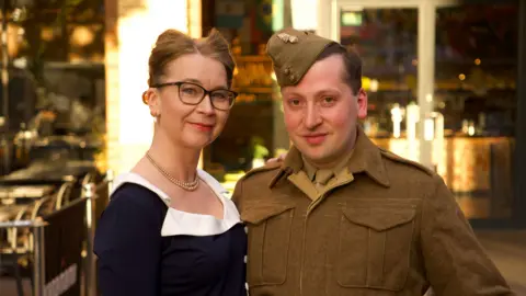 A woman in a blue dress with white collar, pearl necklace and glasses with a man dressed in green army uniform World War Two