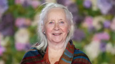 Samir Hussein A woman smiles at the camera in a head and shoulders image. She has shoulder length grey hair swept back from her face and pale blue eyes. She is wearing a terracota coloured shirt and a pashmina shawl in reds, browns and blues over her shoulders