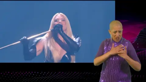 A woman in a purple dress on the right hand side of a TV screen doing sign language. A woman who is singing is the main focus of the picture. She is in the middle and dressed in a leather. She has long blonde hair and holding a microphone.