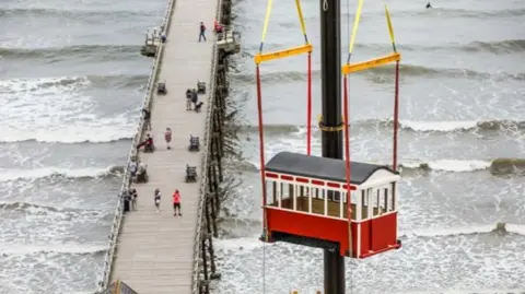 A red and white wooden carriage with a sloped roof being lifted by crane over a pier and sea. People are on the pier and the sea has waves.