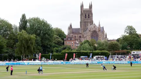 Worcester New Road cricket ground. Players run across the ground which is set just in front of Worcester Cathedral. It is an overcast day and the ground is surrounded by trees and greenery. There are crowds gathered in the stands to watch the game. 