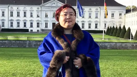 Qian Zhimin - wearing glasses and her hair dyed red - standing in front of a building in Berlin. She is wearing a fur-lined bright blue coat.