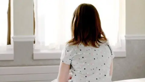 Getty Images A girl with mid length brown hair sits on a bed facing a window. She is wearing a grey top with black, pink and white dots on it