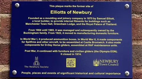 A blue plaque that says: This plaque marks the former site of Elliotts of Newbury." And lists its history in four paragraphs, with the Newbury Society and Newbury Town Council's logos at the bottom, with a QR code in the middle of them.