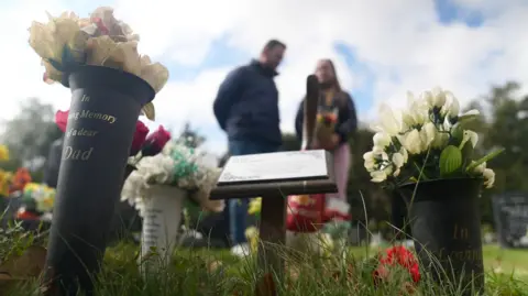 John Fairhall/BBC In the foreground of the picture are several pots of flowers, one with the inscription "In loving memory of a dear dad". In the background a man and woman stand with a bouquet of flowers.