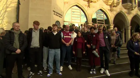 Kate Bradbrook/BBC A group of people are standing in front of the Guildhall building in Northampton. Some of them have their heads bowed.
