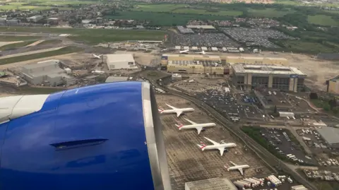 View from a plane shows blue engine of plane and four aircraft on tarmac outside low-rise terminal buildings.