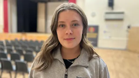 Yvie has long fair hair and his wearing a zipped grey top. She is standing in a hall inside the school.