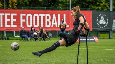 PDR Photos A woman with an amputated leg kicks a ball - she is leaning on her crutches while her body is swinging upwards into the air. She wears a football kit comprised of a black t-shirt, black shorts, and a long black sock and trainer on one leg. She is on a football pitch and three people in chairs and one person standing up watch her in the background.