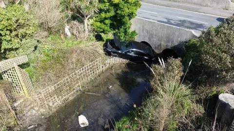 Dan Jessup An aerial photograph shows the black car in a ditch with the road behind it. The car is in water and a fence on the bank is broken.
