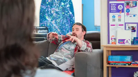 Simon is sitting on a sofa. He has a pair of sunglasses on his forehead. He is wearing a red, white and black check shirt over a t-shirt with a stylised New York skyline on it.