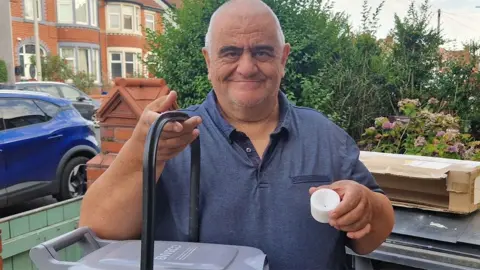 LDRS Brian Roberts, who has white buzzcut hair and a blue polo shirt, stands outdoors in a residential front garden, holding the handle of a grey wheeled bin or trolley in one hand and a small white plastic cup in the other. Behind the person are green bushes, cardboard boxes, a parked blue car, and red-brick houses in daylight.