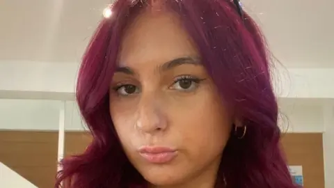 ABC Alicia Kemp, looking at camera, with a pout. She has long hair dyed a reddish purple colour. 