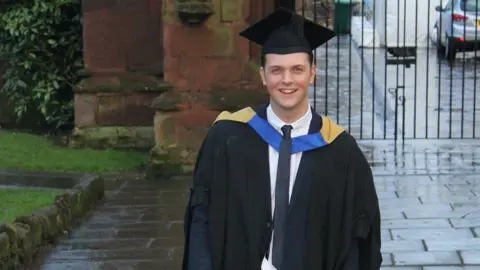 Family photo Jack Hurn, smiling at the camera, he is wearing black university graduation robes and a hat and is standing in front of a gate