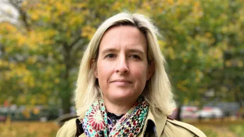 Gemma Lindfield, a woman with short blonde hair wearing a multicoloured patterned scarf and beige trench coat, stood in front of some trees