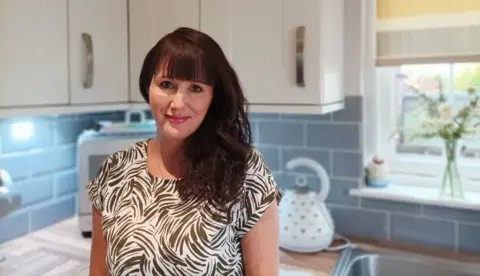 Angela Gates stands in a kitchen smiling and wearing a patterned top