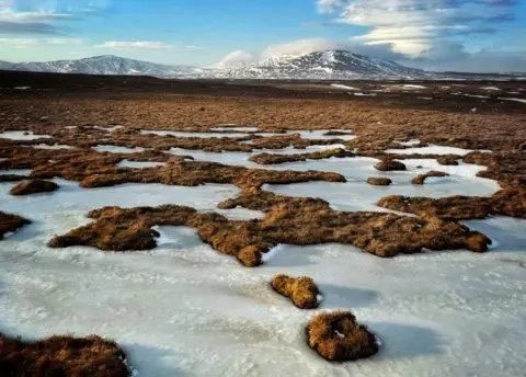 Peter Xu Patchy frozen ground stretches across a wide moorland, with brown grasses breaking through the ice. Snow‑covered mountains rise in the background beneath a bright blue sky with scattered clouds.