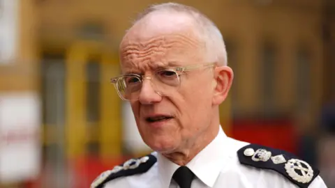 PA Media Close-up profile of Sir Mark Rowley wearing a white uniform shirt, black tie, and detailed epaulettes against a blurred urban background.