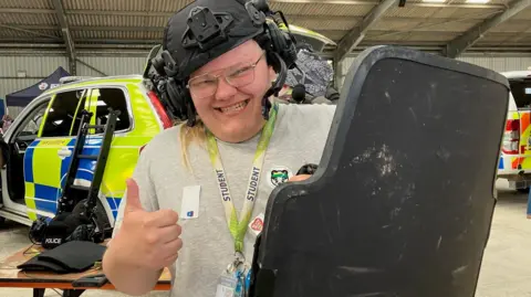 Louis is wearing a heavy-duty police helmet which has built in headphones and a camera mount at the front. He is holding a riot shield up and is smiling and giving a thumbs up to the camera. He is wearing a lanyard that says he is a student, he has blond hair and is wearing a pair of glasses. Behind him are two police cars.
