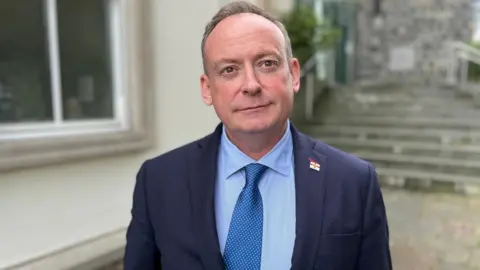 Deputy Lyndon Trott wearing a blue suit, blue shirt, blue metallic tie wearing a Guernsey flag lapel pin looking towards the camera. 