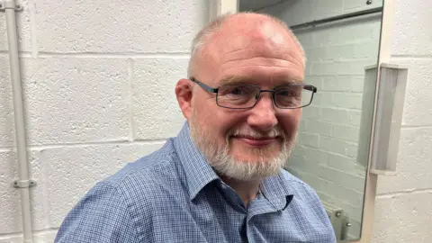 Oliver Conopo/ BBC A man with a bald head and a grey beard with black thin framed glasses on is looking at the camera with a soft smile. He is wearing a light blue checkered shirt and is standing front of a white wall. 