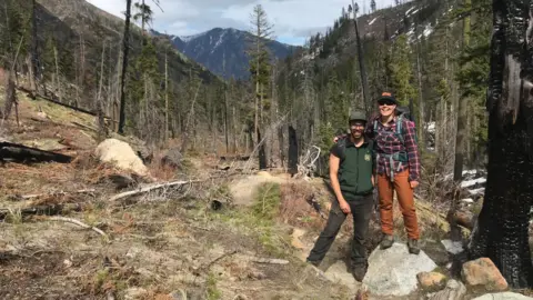 Subjected to BBC Demetrios and Thompson Stand smiling in the middle of a clearing in a hilly forest, with a visible mountain peak in the background. Demetrios has a beard and wears a green sports vest and brown work pants and brown hiking boots, with a baseball cap protecting your eyes. Thompson stands next to him on a rock so that she is larger, wearing orange work pants, a red flannel shirt, a baseball hat and a hiking backpack. 