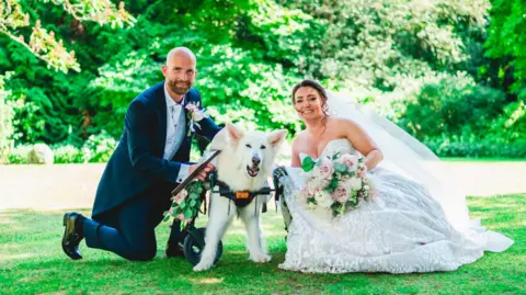 Rich Smith, left, in a navy blue suit and a silver waistcoat. He is bald with a beard. He kneels on the floor and holds his dog, who is white and fluffy and supported by wheels due to being ill. His wife is on the right and wears a lacy dress and veil. She has dark hair and holds a bouquet. 