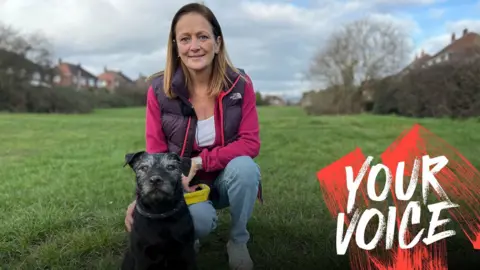 Jim Connolly/BBC Kelly Norton, wearing a pink top and gilet, crouches down in a park with her dog Vinny
