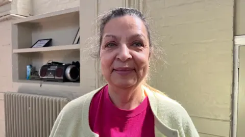 Ellen Knight/BBC Photo of Lesley looking into the camera and smiling slightly. She is wearing a bright pink t-shirt with a light yellow cardigan over the top. She has long dark hair that is tied back into a ponytail. Lesley is pictured stood in front of a brick wall painted light green, with a shelf behind her. On the shelf are some framed certificates, and a stereo. 