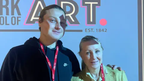 BBC/ Emily Johnson A man and a woman standing in front of a wall, both wearing red lanyard badges, one in a black Nike hoodie and the other in a light blazer, with shadows cast across their faces.