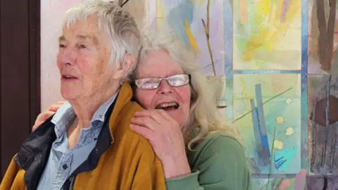 Elspeth Owen Julia Ball and Elspeth Owen in front of a colourful canvas in 2014. Julia is on the left and has short white hair and is wearing an orange jacket over a light blue denim shirt. Elspeth is behind her with long white hair and wearing glasses and a light green jumper. She is leaning on Julia's back and laughing while looking towards the camera.