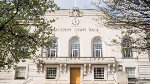 Getty Images The facade of Hackney Town Hall, a cream colored stone building with large arched windows and a clock above the entrance. Steps lead up to the main door and trees frame the picture, partially covering the building.