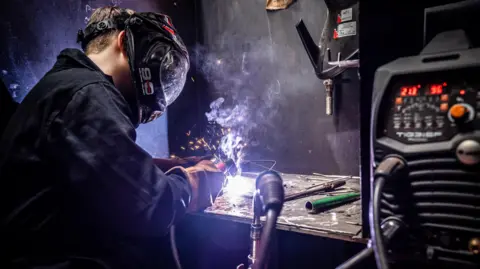 RNN Group A young man dressed in dark overalls, thick brown gloves and a protective mask stands at a work counter in a corner of a room with black walls. He is welding some metal and sparks can be seen. To his right there is a large black piece of equipment with a tube connected to the welding tool.