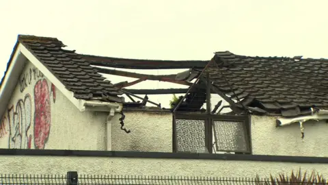 The roof of a building after a fire. Large parts of the roof is missing. The sky is grey and cloudy. Old graffiti is on the side of the building. 