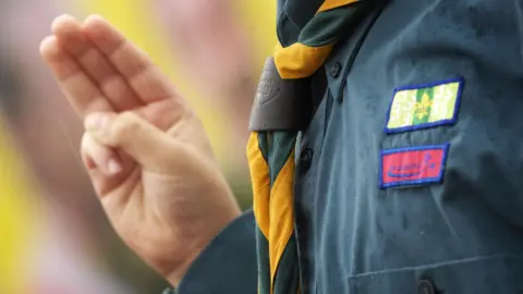 A close-up of a scout wearing a dark green shirt and a yellow scarf, who is holding up a hand in a three-finger salute.
