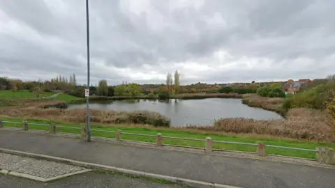 Oakley Vale Lake, showing a large lake with water, a road in front, with a low railing, a post, and trees in the distance. There are reeds around the lake and houses in the distance to the right and at the back of the image. 