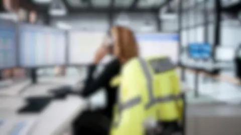 A view of a blurred emergency services call room, there is a woman sat in front of a desk with a phone to her ear